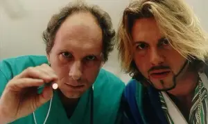 Dwóch mężczyzn, jeden w zielonym kitlu trzymający mały przedmiot, drugi z blond włosami i złotym łańcuchem. Scena z filmu "Chłopaki nie płaczą 2".