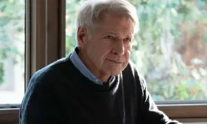 Harrison Ford, z siwymi włosami i zmęczonym spojrzeniem, siedzi przy oknie, ubrany w sweter i koszulę.