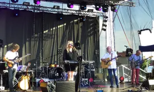 Zespół gra na scenie, wokalistka przy klawiszach, gitarzyści i basista. W tle perkusja. To może być koncert promujący nowe tytuły albumów grupy pod budą.