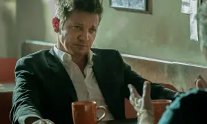 Burmistrz Kingstown, Jeremy Renner, w garniturze i rozpiętej koszuli, siedzi przy stole z kubkiem kawy, w zamyśleniu słuchając rozmówcy.