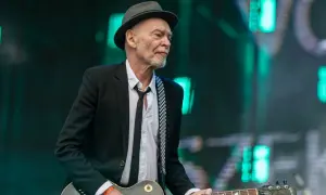 Charyzmatyczny gitarzysta w kapeluszu i czarnej marynarce, z rozpiętą białą koszulą i czarnym krawatem, gra na gitarze Gibson Les Paul.