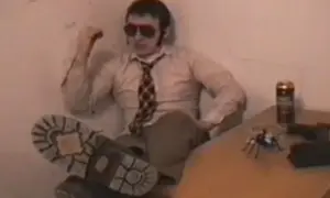 Facet w okularach i krawacie, z nogą na stole, obok puszka i zabawkowego pająka. Wygląda jakby miał zaraz zacząć opowiadać historie o "kurach z grubej rury".