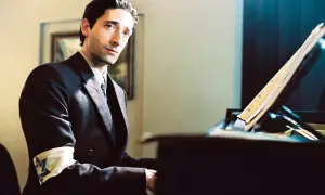Adrien Brody jako Władysław Szpilman w filmie "Pianista" Romana Polańskiego, z opaską na ramieniu, siedzi przy fortepianie.