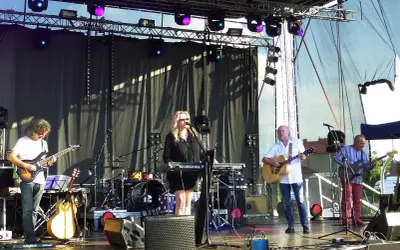 Zespół gra na scenie, wokalistka przy klawiszach, gitarzyści i basista. W tle perkusja. To może być koncert promujący nowe tytuły albumów grupy pod budą.