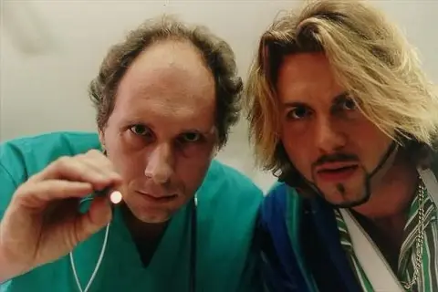 Dwóch mężczyzn, jeden w zielonym kitlu trzymający mały przedmiot, drugi z blond włosami i złotym łańcuchem. Scena z filmu "Chłopaki nie płaczą 2".