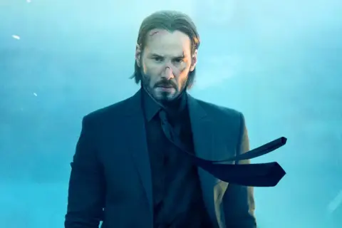 John Wick, z bliznami na twarzy, w garniturze, czeka na premierę John Wick 5.