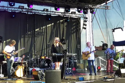 Zespół gra na scenie, wokalistka przy klawiszach, gitarzyści i basista. W tle perkusja. To może być koncert promujący nowe tytuły albumów grupy pod budą.