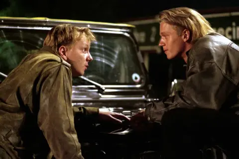 Dwóch mężczyzn w skórzanych kurtkach pochyla się nad maską samochodu. Scena z filmu o mafii, pełna napięcia.