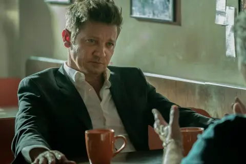 Burmistrz Kingstown, Jeremy Renner, w garniturze i rozpiętej koszuli, siedzi przy stole z kubkiem kawy, w zamyśleniu słuchając rozmówcy.