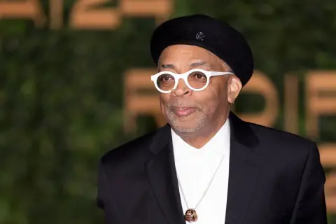 Spike Lee w czarnym beretce i białych okularach, od góry do dołu w czarnym garniturze i białej koszuli.