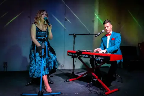 Wokalistka w niebieskiej sukience i pianista w garniturze tworzą duet. Ich występ to przykład kameralnych zespołów wokalnych.