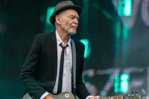 Charyzmatyczny gitarzysta w kapeluszu i czarnej marynarce, z rozpiętą białą koszulą i czarnym krawatem, gra na gitarze Gibson Les Paul.
