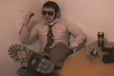 Facet w okularach i krawacie, z nogą na stole, obok puszka i zabawkowego pająka. Wygląda jakby miał zaraz zacząć opowiadać historie o "kurach z grubej rury".
