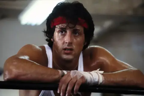 Spocony Rocky Balboa, z czerwoną opaską na głowie, opiera się o ring.