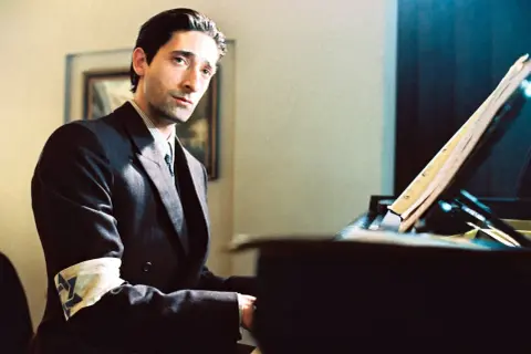Adrien Brody jako Władysław Szpilman w filmie "Pianista" Romana Polańskiego, z opaską na ramieniu, siedzi przy fortepianie.