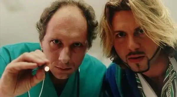 Dwóch mężczyzn, jeden w zielonym kitlu trzymający mały przedmiot, drugi z blond włosami i złotym łańcuchem. Scena z filmu "Chłopaki nie płaczą 2".