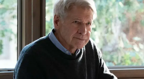 Harrison Ford, z siwymi włosami i zmęczonym spojrzeniem, siedzi przy oknie, ubrany w sweter i koszulę.