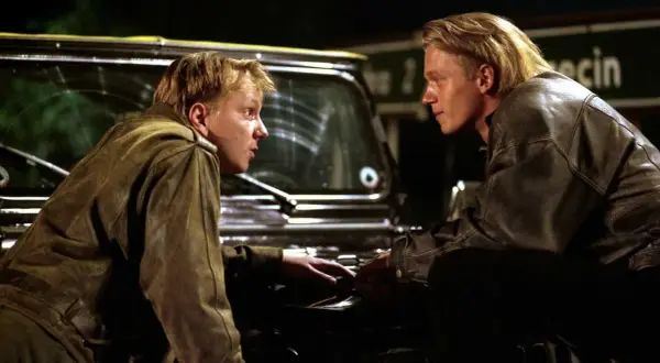 Dwóch mężczyzn w skórzanych kurtkach pochyla się nad maską samochodu. Scena z filmu o mafii, pełna napięcia.