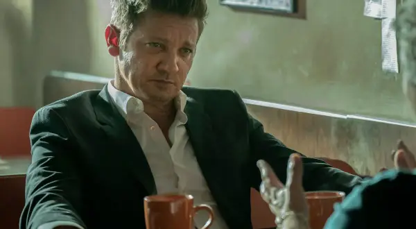 Burmistrz Kingstown, Jeremy Renner, w garniturze i rozpiętej koszuli, siedzi przy stole z kubkiem kawy, w zamyśleniu słuchając rozmówcy.