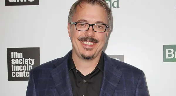 Vince Gilligan, twórca filmów, seriali i programów, w garniturze i okularach, uśmiecha się na tle logo "Film Society Lincoln Center" i symbolu pierwiastka "Br".