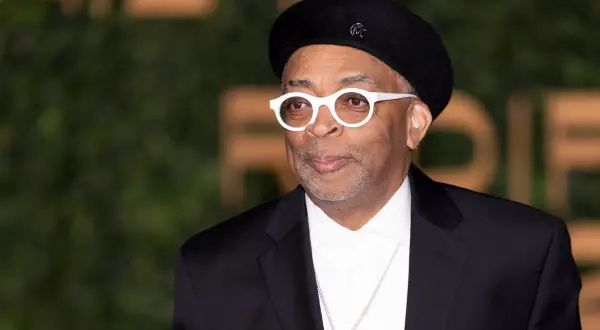 Spike Lee w czarnym beretce i białych okularach, od góry do dołu w czarnym garniturze i białej koszuli.