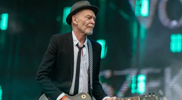 Charyzmatyczny gitarzysta w kapeluszu i czarnej marynarce, z rozpiętą białą koszulą i czarnym krawatem, gra na gitarze Gibson Les Paul.
