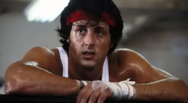 Spocony Rocky Balboa, z czerwoną opaską na głowie, opiera się o ring.