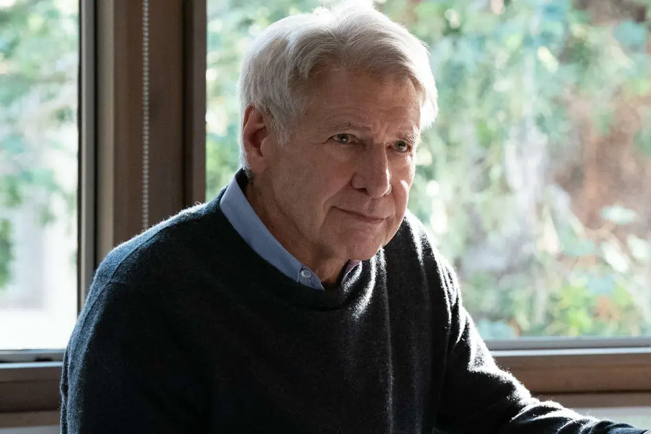Harrison Ford, z siwymi włosami i zmęczonym spojrzeniem, siedzi przy oknie, ubrany w sweter i koszulę.