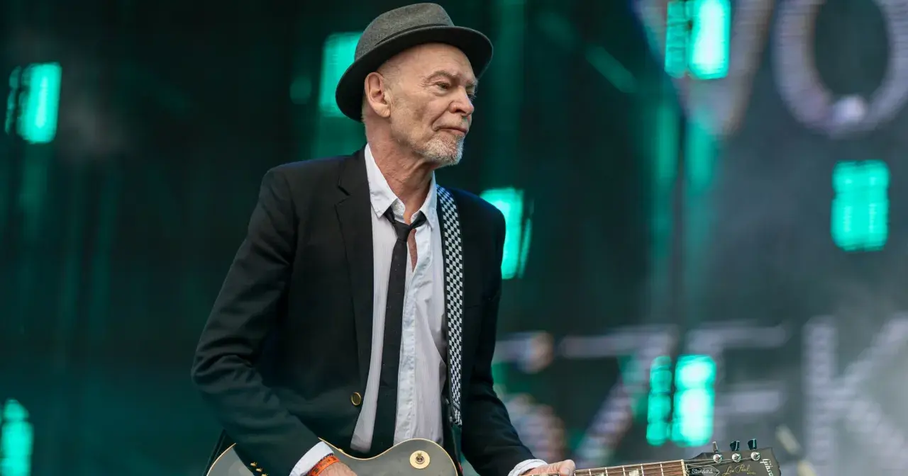 Charyzmatyczny gitarzysta w kapeluszu i czarnej marynarce, z rozpiętą białą koszulą i czarnym krawatem, gra na gitarze Gibson Les Paul.