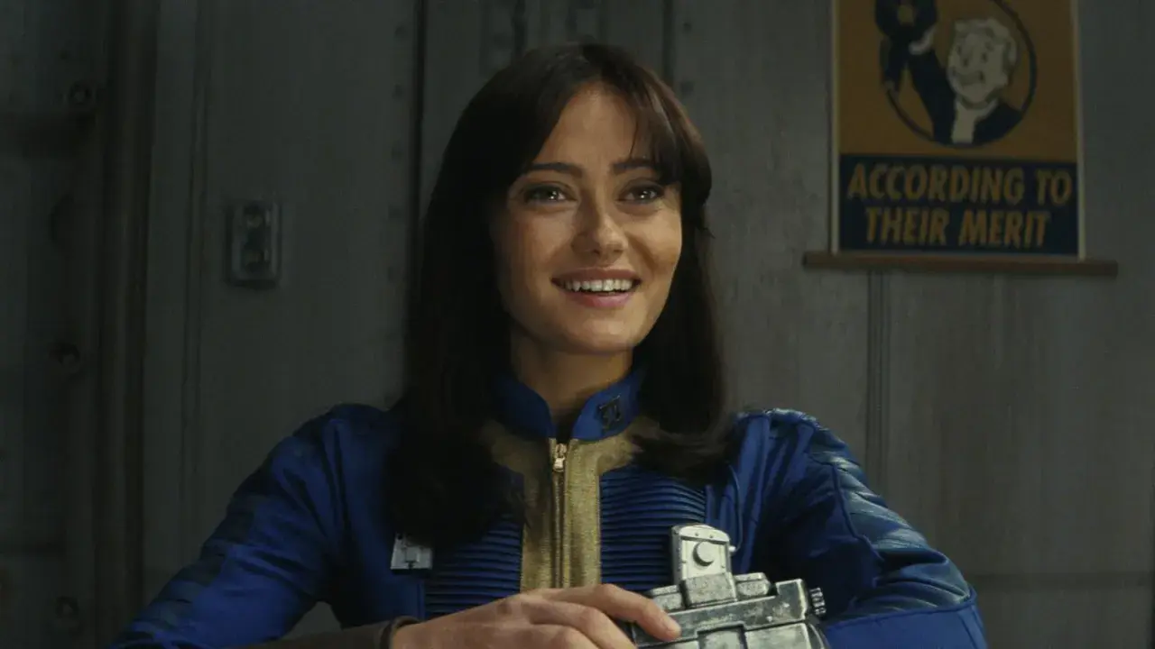Uśmiechnięta Lucy z serialu Fallout, w niebieskim kombinezonie, trzymająca urządzenie. Na ścianie wisi plakat "According to their merit".