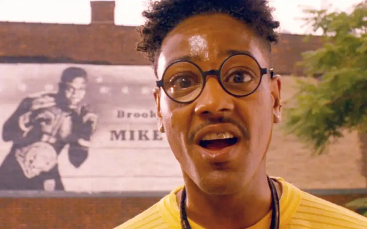 Giancarlo Esposito w okularach i żółtej koszulce, z plakatem boksera w tle.