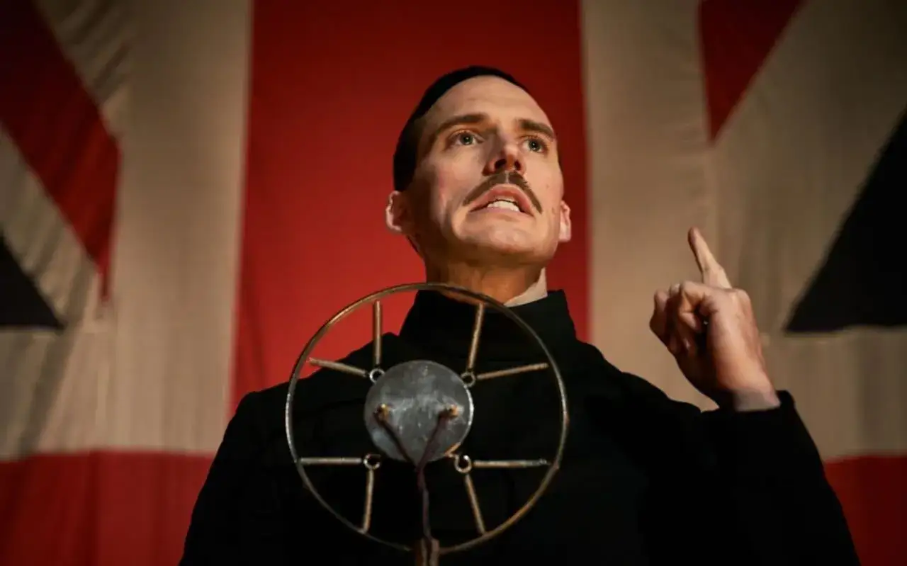 Oswald Mosley przemawia do tłumu na tle flagi w Peaky Blinders sezon 5. Jego gest i wyraz twarzy sugerują płomienne przemówienie.