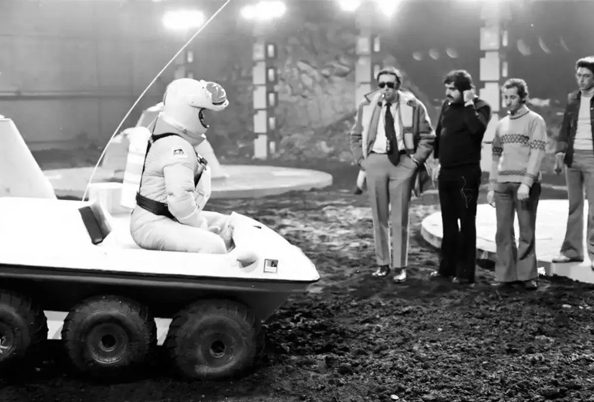 Astronaut w skafandrze siedzi w pojeździe, podczas gdy ekipa