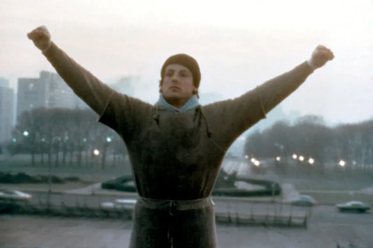 Rocky Balboa, w czapce i dresie, wznosi ręce w geście triumfu na tle miejskiego krajobrazu.