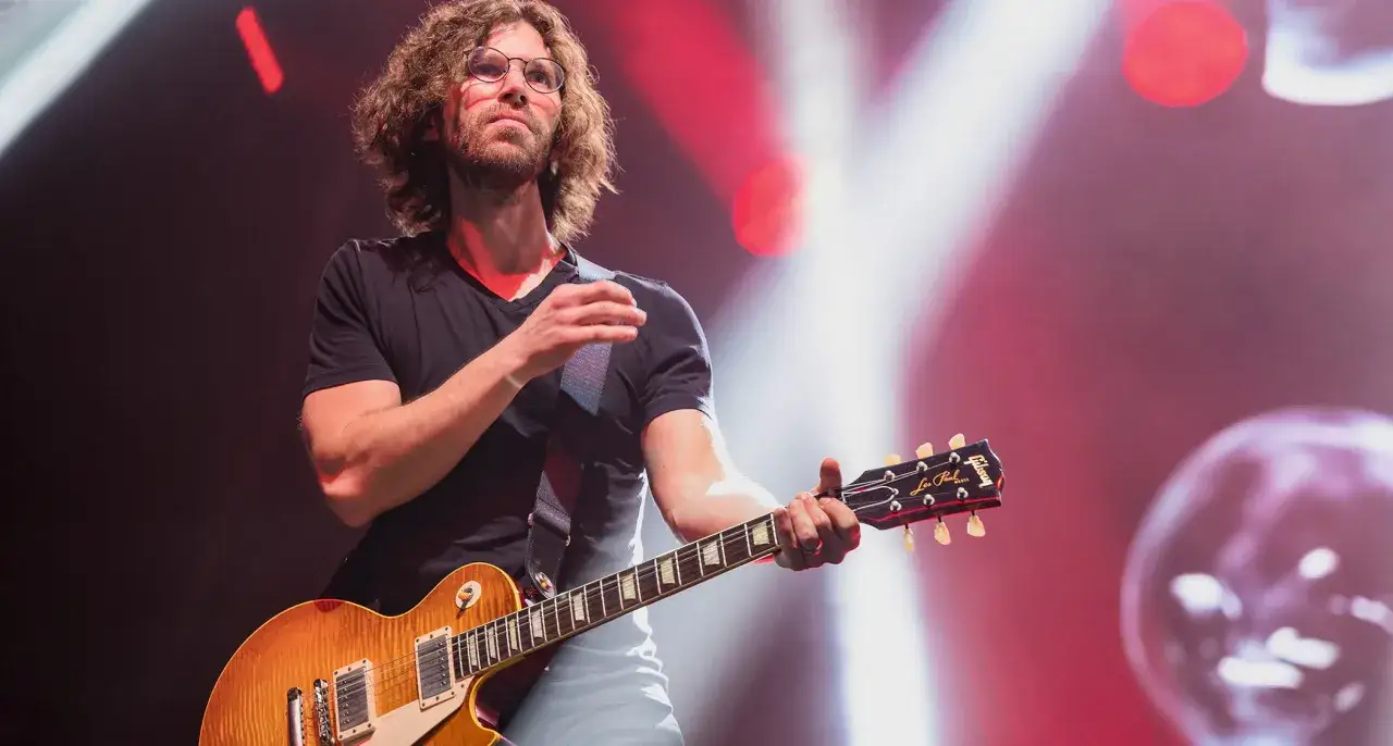 Charyzmatyczny gitarzysta w okularach z kręconymi włosami gra na gitarze Gibson Les Paul.