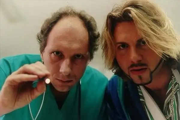 Dwóch mężczyzn, jeden w zielonym kitlu trzymający mały przedmiot, drugi z blond włosami i złotym łańcuchem. Scena z filmu "Chłopaki nie płaczą 2".