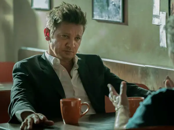 Burmistrz Kingstown, Jeremy Renner, w garniturze i rozpiętej koszuli, siedzi przy stole z kubkiem kawy, w zamyśleniu słuchając rozmówcy.