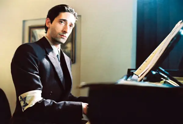 Adrien Brody jako Władysław Szpilman w filmie "Pianista" Romana Polańskiego, z opaską na ramieniu, siedzi przy fortepianie.